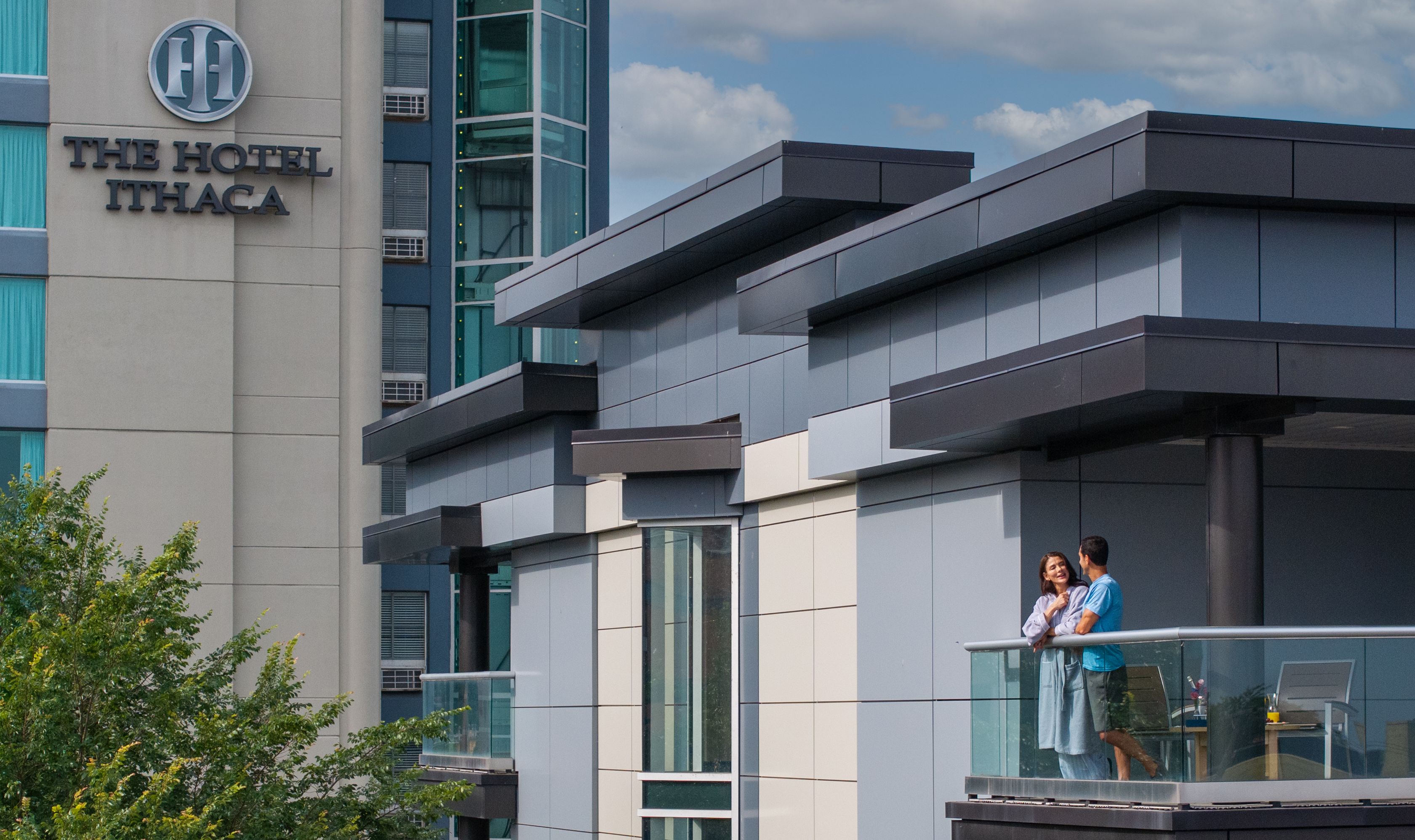 A couple steps out onto the balcony of a room at The Hotel Ithaca, with the hotel logo visible in the background